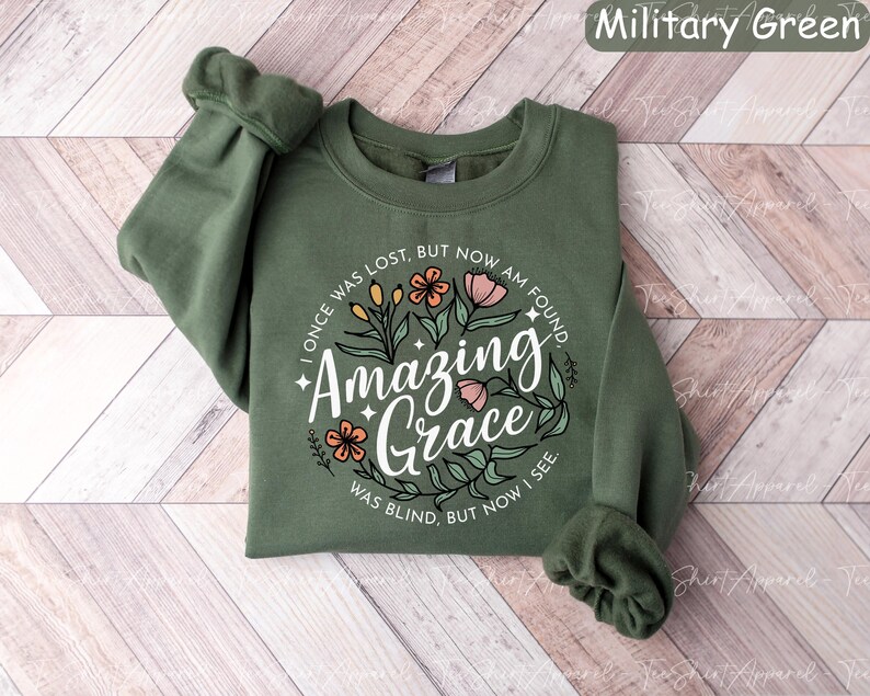 May include: A military green sweatshirt with the text "Amazing Grace" in a floral design. The text "I once was lost, but now am found, was blind, but now I see" is also included. The sweatshirt has long sleeves.