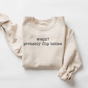 Peut inclure: Un sweat-shirt cr&egrave;me avec le texte "wwjd? probably flip tables" en noir. Les manches longues sont retrouss&eacute;es. Le sweat-shirt est fait d'une mati&egrave;re douce.