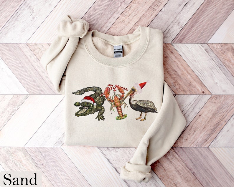 May include: Sand-colored sweatshirt featuring a festive design of an alligator, lobster, and pelican, each wearing a Santa hat. The alligator is green, the lobster is red, and the pelican is gray. The sweatshirt has a crew neck and long sleeves.