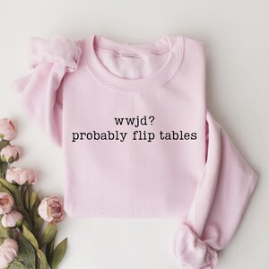 Peut inclure: Un sweat-shirt rose clair avec le texte "wwjd? probably flip tables" en noir. Le sweat-shirt est pli&eacute; et entour&eacute; de fleurs roses et de feuilles vertes.