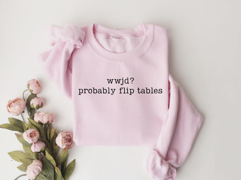 Peut inclure: Un sweat-shirt rose clair avec le texte "wwjd? probably flip tables" en noir. Le sweat-shirt est pli&eacute; et entour&eacute; de fleurs roses et de feuilles vertes.