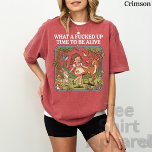May include: Crimson-colored t-shirt with the text "WHAT A FUCKED UP TIME TO BE ALIVE." The graphic features a cartoon illustration of a girl, deer, rabbit, and butterflies in a forest setting. The shirt is a casual, oversized fit.