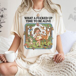 May include: Ivory-colored t-shirt with the text "WHAT A FUCKED UP TIME TO BE ALIVE." The graphic features a cartoon girl, deer, rabbit, and butterflies in a forest scene. The shirt is a casual, comfortable style.