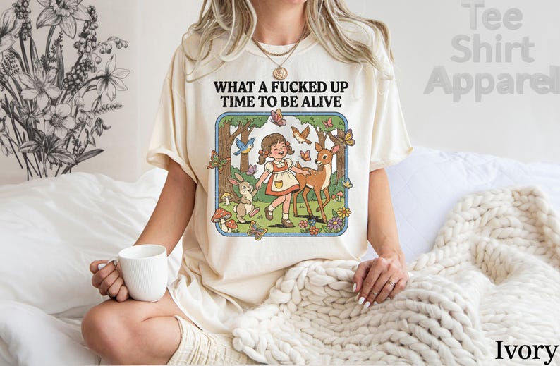 May include: Ivory-colored t-shirt with the text "WHAT A FUCKED UP TIME TO BE ALIVE." The graphic features a cartoon girl, deer, rabbit, and butterflies in a forest scene. The shirt is a casual, comfortable style.