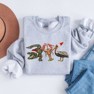 May include: A light gray sweatshirt featuring a festive design with an alligator, lobster, and pelican, each wearing a Santa hat. The alligator is green, the lobster is red, and the pelican is gray. The sweatshirt is paired with blue jeans and a brown hat.