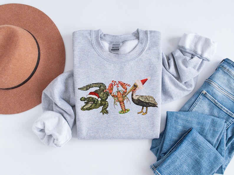 May include: A light gray sweatshirt featuring a festive design with an alligator, lobster, and pelican, each wearing a Santa hat. The alligator is green, the lobster is red, and the pelican is gray. The sweatshirt is paired with blue jeans and a brown hat.