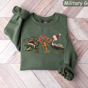 May include: Military green sweatshirt featuring a festive design. The graphic includes a green alligator, a red crawfish, and a pelican, all wearing Santa hats. The text "Military Green" is in the upper right corner.