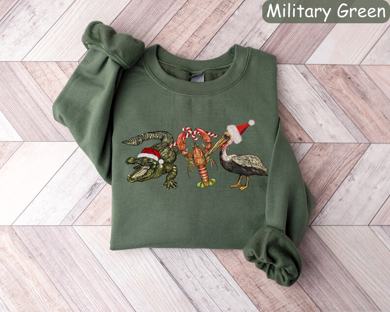May include: Military green sweatshirt featuring a festive design. The graphic includes a green alligator, a red crawfish, and a pelican, all wearing Santa hats. The text "Military Green" is in the upper right corner.