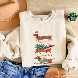 May include: Cream-colored sweatshirt featuring three dachshunds in festive attire. The top dachshund wears a Santa hat and scarf, the middle one carries a Christmas tree, and the bottom one skis. Text reads "Dachshund Through The Snow."