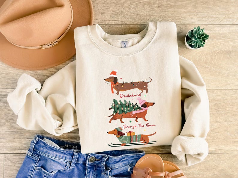 May include: Cream-colored sweatshirt featuring three dachshunds in festive attire. The top dachshund wears a Santa hat and scarf, the middle one carries a Christmas tree, and the bottom one skis. Text reads "Dachshund Through The Snow."
