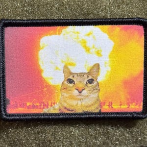Peut inclure: Un écusson rectangulaire à bordure noire cousue représentant un chat tigré brun centré devant une grande explosion en forme de champignon blanche et orange sur un fond vibrant aux tons rouges et jaunes.