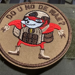 Könnte beinhalten: Ein brauner gestickter Aufnäher mit einer roten Comicfigur, die einen Helm trägt und eine Waffe hält. Der Text "DO U NO DE WAE?" ist um die Figur herum geschrieben.