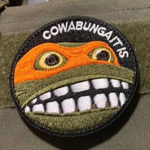 May include: Round embroidered patch with a cartoon turtle face, orange mask, and a wide grin. The text "COWABUNGAITIS" is in white on a black background. The patch has a green and black border and is likely for clothing or accessories.