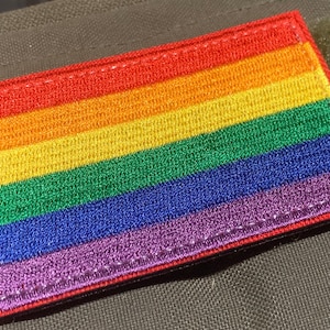 May include: A rainbow pride flag patch with red, orange, yellow, green, blue, and purple stripes. The patch is embroidered and has a red stitched border.