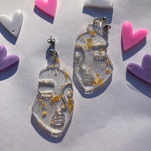 May include: A pair of clear resin earrings with gold leaf accents. The earrings are shaped like a face with a simple outline and feature a gold leaf design.