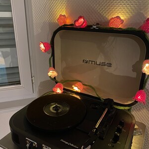 May include: A black vintage-style record player with a black vinyl record on the turntable. The record player has a silver handle and the words "Vintage Collection" printed on the front. A string of pink crocheted flower lights is wrapped around the record player.
