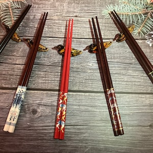 May include: Five pairs of chopsticks with decorative designs. The chopsticks are made of wood and have a variety of colors and patterns. The chopsticks are arranged in a row on a wooden surface.