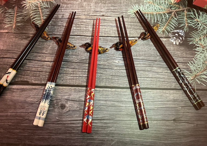 May include: Five pairs of chopsticks with decorative designs. The chopsticks are made of wood and have a variety of colors and patterns. The chopsticks are arranged in a row on a wooden surface.