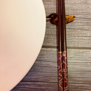 May include: A pair of brown chopsticks with a red and blue dragon design rest in a duck-shaped chopstick holder on a wooden surface. A white plate is to the left of the chopsticks.