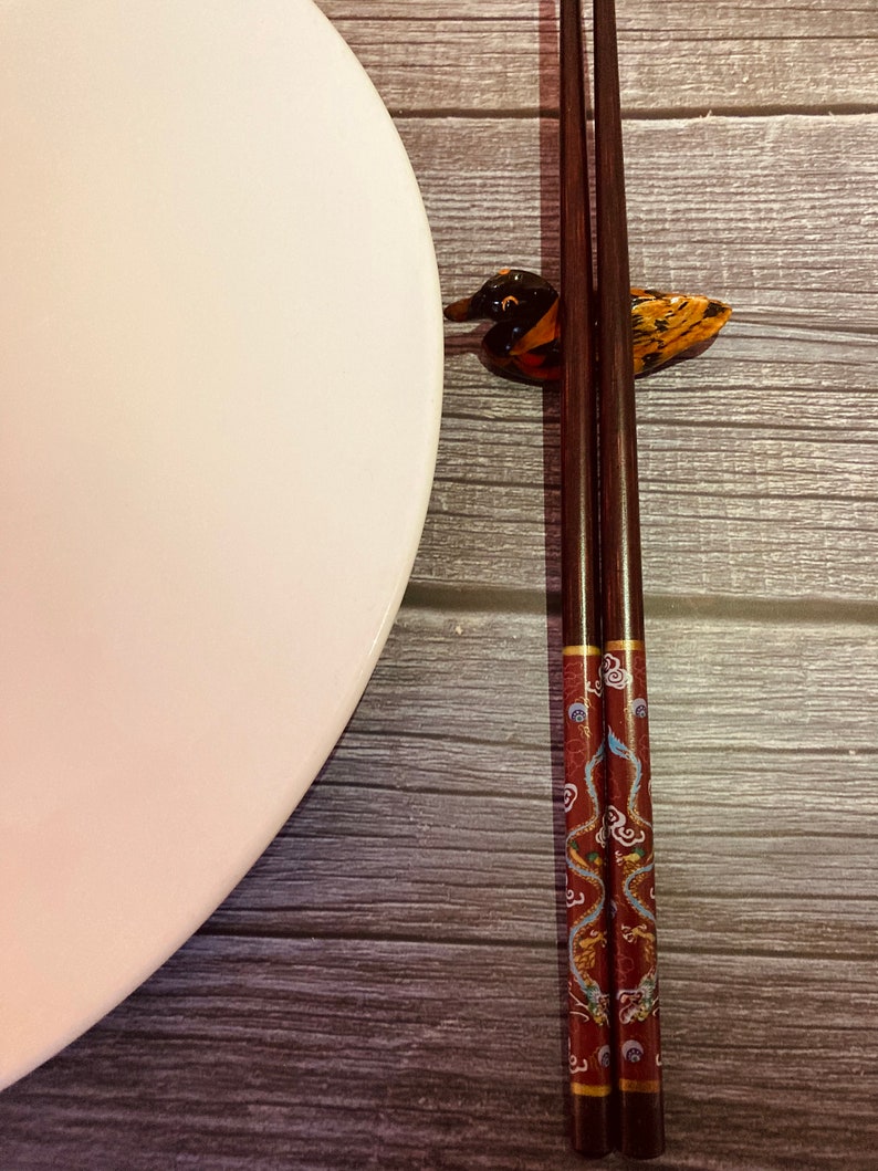 May include: A pair of brown chopsticks with a red and blue dragon design rest in a duck-shaped chopstick holder on a wooden surface. A white plate is to the left of the chopsticks.