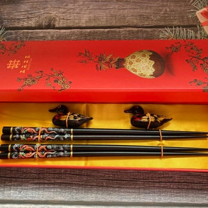 May include: A red box with a floral design and the Chinese character for double happiness. The box contains a set of black chopsticks with gold and red accents and two decorative duck figurines.
