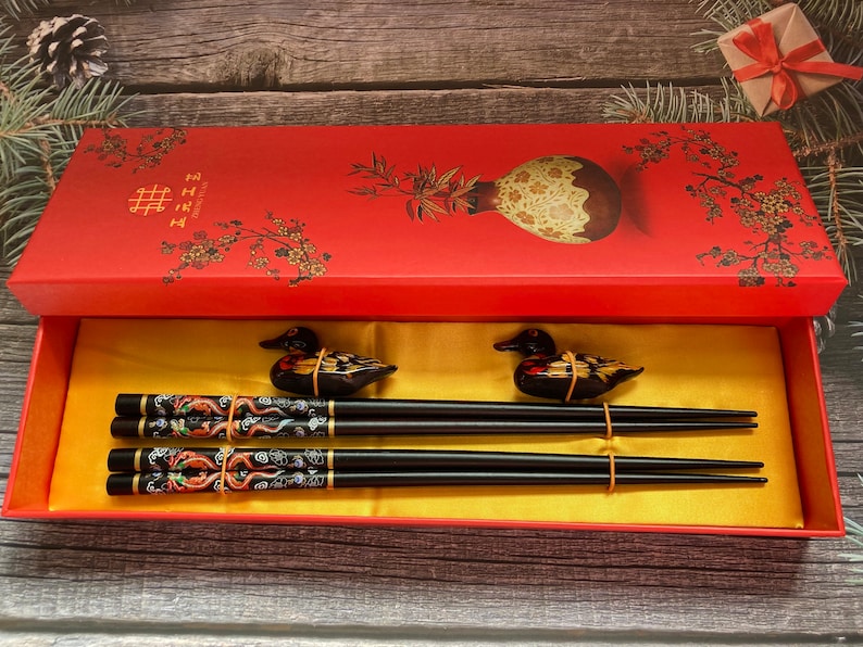 May include: A red box with a floral design and the Chinese character for double happiness. The box contains a set of black chopsticks with gold and red accents and two decorative duck figurines.