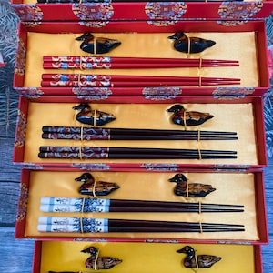 May include: Five sets of chopsticks in red and black boxes with duck-shaped handles. Each set includes two chopsticks with a decorative design. The boxes have a gold background with Chinese characters.