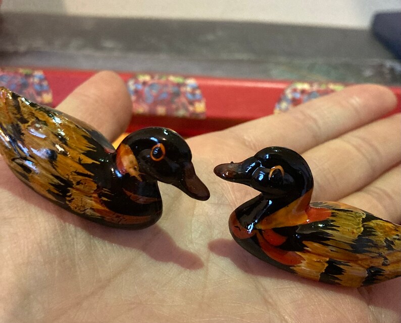 May include: Two small wooden duck figurines with black, brown, and orange paint. The ducks are facing different directions and have detailed features.