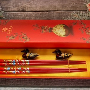 May include: A red box with gold Chinese characters and a floral design contains a set of red chopsticks with gold accents and two black and gold duck figurines.