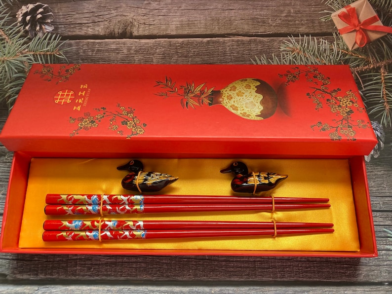 May include: A red box with gold Chinese characters and a floral design contains a set of red chopsticks with gold accents and two black and gold duck figurines.