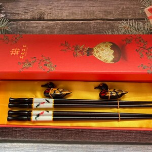 May include: A red box with a gold interior and a floral design. The box contains two pairs of black chopsticks with duck-shaped handles. The chopsticks are decorated with a white and gold design. The box has the Chinese character for "double happiness" on the lid.