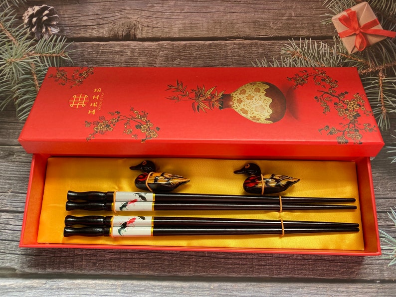 May include: A red box with a gold interior and a floral design. The box contains two pairs of black chopsticks with duck-shaped handles. The chopsticks are decorated with a white and gold design. The box has the Chinese character for "double happiness" on the lid.