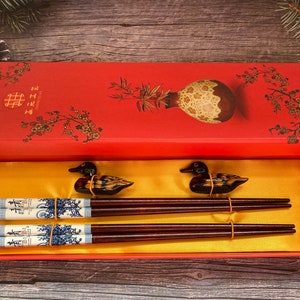 May include: A red box with a floral design and the text "囍" on the lid. The box is open and contains two pairs of chopsticks with duck-shaped handles. The chopsticks are decorated with a blue and white pattern.