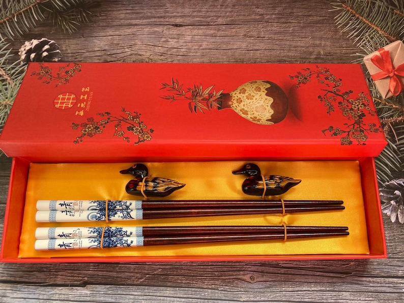 May include: A red box with a floral design and the text "囍" on the lid. The box is open and contains two pairs of chopsticks with duck-shaped handles. The chopsticks are decorated with a blue and white pattern.