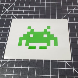 May include: Green pixelated alien spaceship, a classic video game character, on a white background.