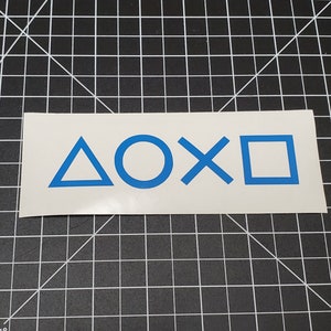 May include: A white sticker featuring the blue PlayStation controller button symbols: triangle, circle, X, and square. The sticker is rectangular and placed on a black cutting mat with a grid pattern.