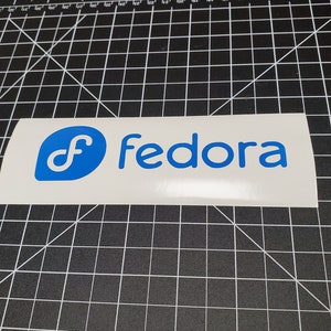 May include: A white sticker with a blue Fedora logo and the word "Fedora" in blue text.