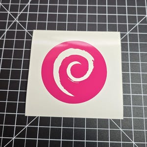 May include: A square sticker with a white background featuring a pink circle with a white spiral design in the center. The sticker is placed on a black and white grid surface.