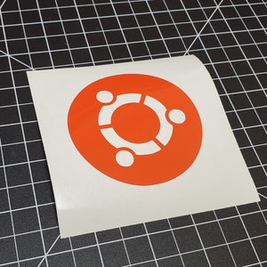 May include: A white sticker with an orange circle containing a white stylized logo of three figures forming a circle. The logo is the symbol for the Linux operating system.