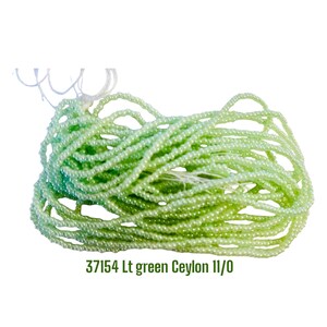 May include: A collection of light green Ceylon 11/0 seed beads strung on white thread. The beads have a smooth, pearlescent finish and are arranged in multiple loops. The text 37154 Lt green Ceylon 11/0 is printed in green below the beads.