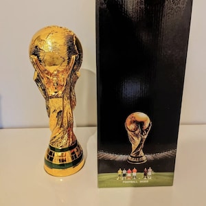 May include: A gold-colored replica of the FIFA World Cup trophy. The trophy is a stylized globe with two figures holding it up. The trophy is on a white surface next to a black box with a picture of the trophy and the words 'FIFA World Cup' and 'Football 2022'.
