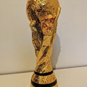 May include: A gold-colored trophy with a globe on top. The trophy is shaped like a person holding up the globe. The base of the trophy has a black band with the word 'FIFA' written on it.