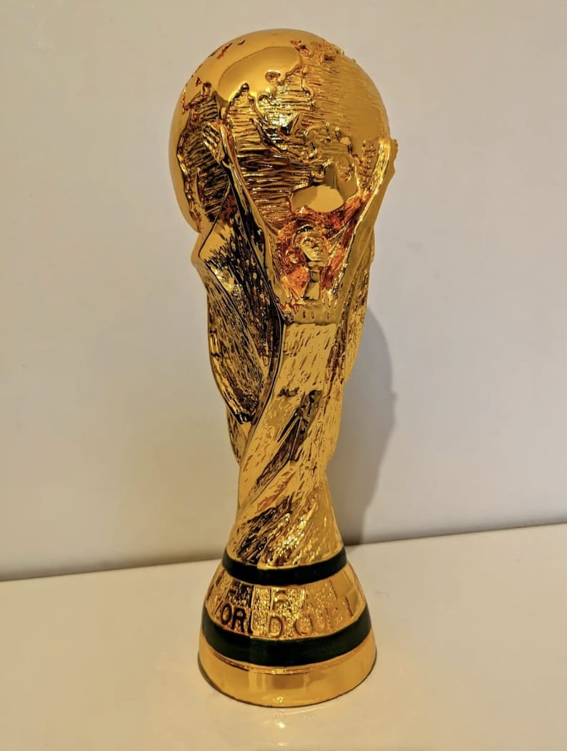 May include: A gold-colored trophy with a globe on top. The trophy is shaped like a person holding up the globe. The base of the trophy has a black band with the word 'FIFA' written on it.