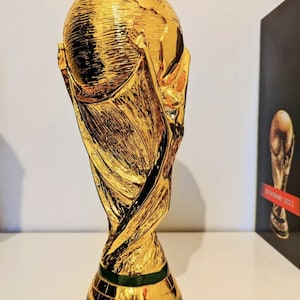 May include: A gold-colored replica of the FIFA World Cup trophy. The trophy is a stylized representation of two human figures holding up the globe. The base of the trophy is green and has a textured surface.