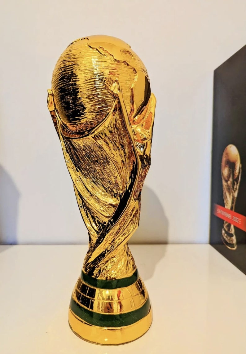 May include: A gold-colored replica of the FIFA World Cup trophy. The trophy is a stylized representation of two human figures holding up the globe. The base of the trophy is green and has a textured surface.