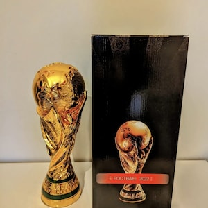 May include: A gold-colored replica of the FIFA World Cup trophy. The trophy is on a white surface next to a black box with a gold-colored image of the trophy on the front. The box has the text 'FIFA FOOTBALL 2022' on the front.