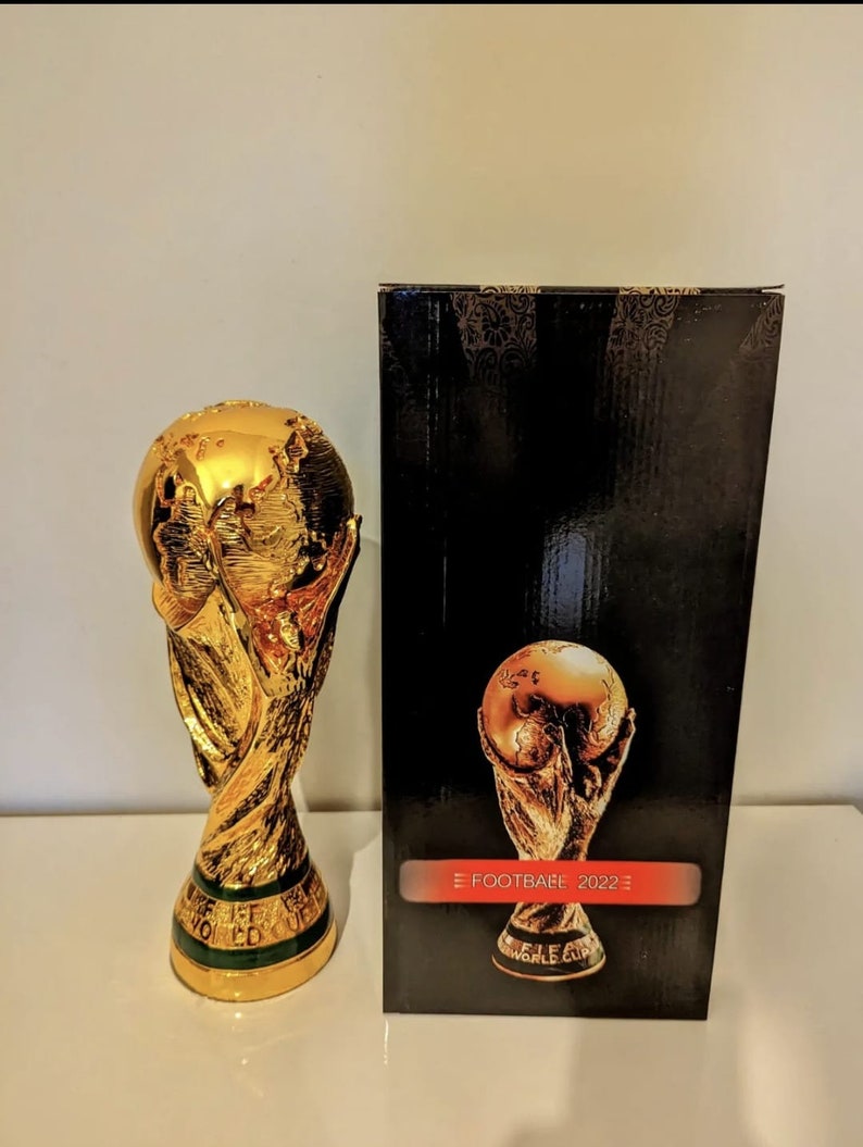 May include: A gold-colored replica of the FIFA World Cup trophy. The trophy is on a white surface next to a black box with a gold-colored image of the trophy on the front. The box has the text 'FIFA FOOTBALL 2022' on the front.