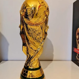 May include: A gold-colored trophy with a globe on top and a green base. The trophy is shaped like a person holding up the globe. The trophy is a replica of the FIFA World Cup trophy.
