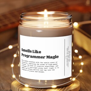 May include: A brown glass candle with a white label that reads "Smells Like Programmer Magic". The candle is lit and the flame is visible. The candle is sitting on a wooden coaster and there are white fairy lights in the background.