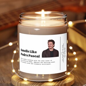 May include: A lit candle in a glass jar with a label that reads "Smells Like Pedro Pascal." The label features a photo of Pedro Pascal. The candle sits on a wooden coaster, with string lights and a guitar in the background.